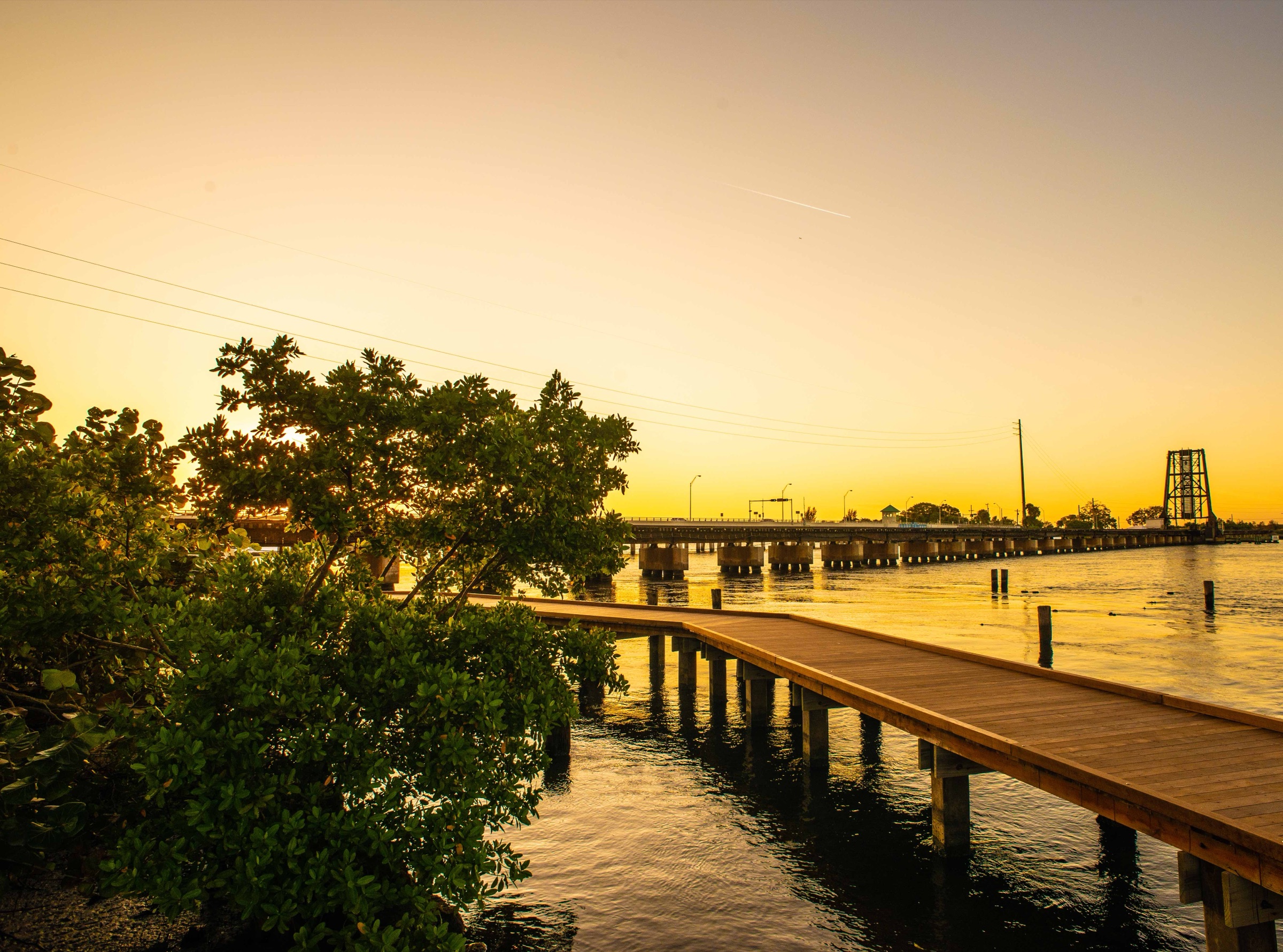 A wooden boardwalk over the St. Lucie River in Stuart at sunset, with the FEC railroad bridge in the distance and mangroves at the left.