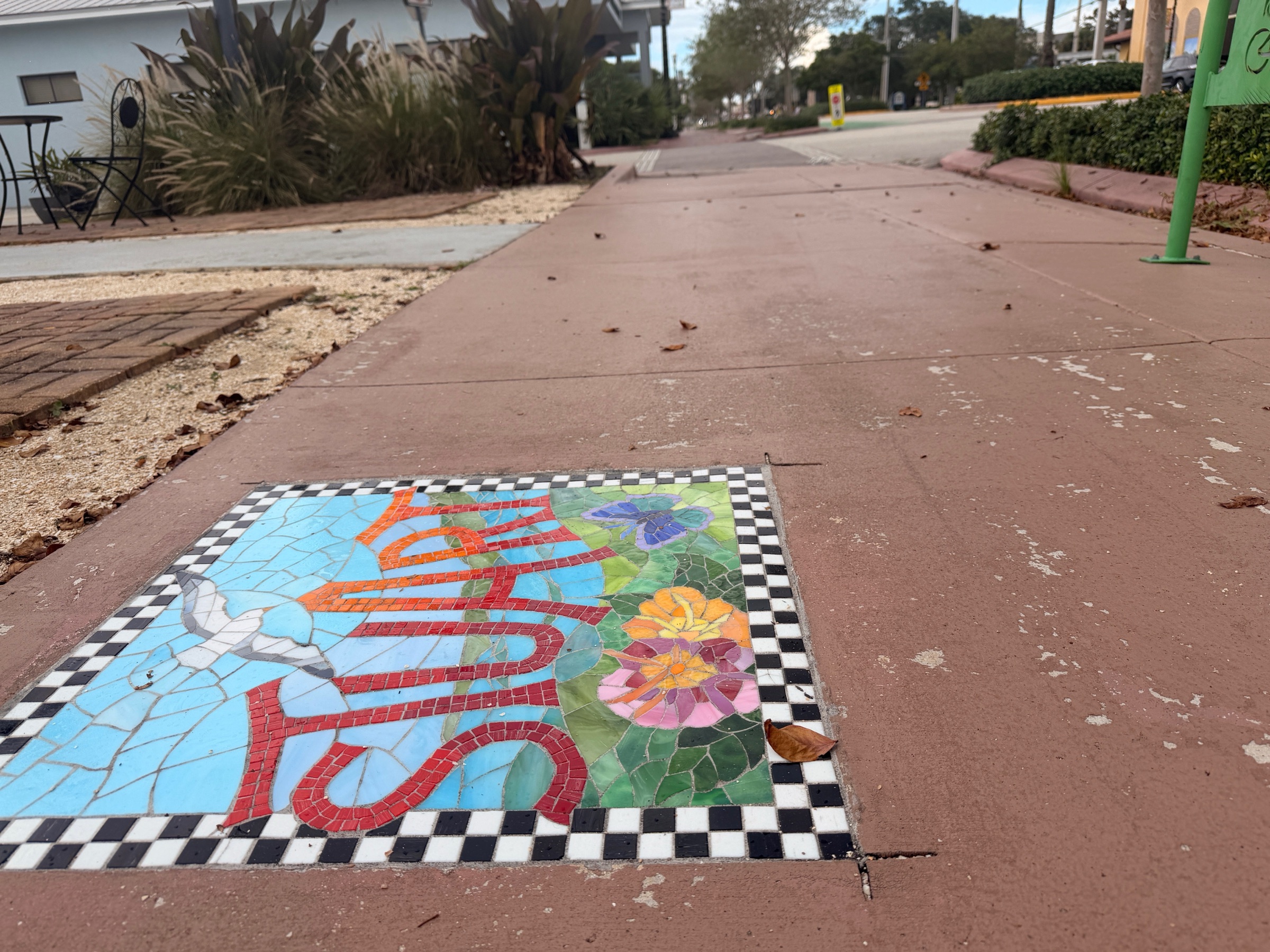 A stained-glass sidewalk mosaic set into a terracotta sidewalk in downtown Stuart, with storefronts and a cafe table further down the walk.